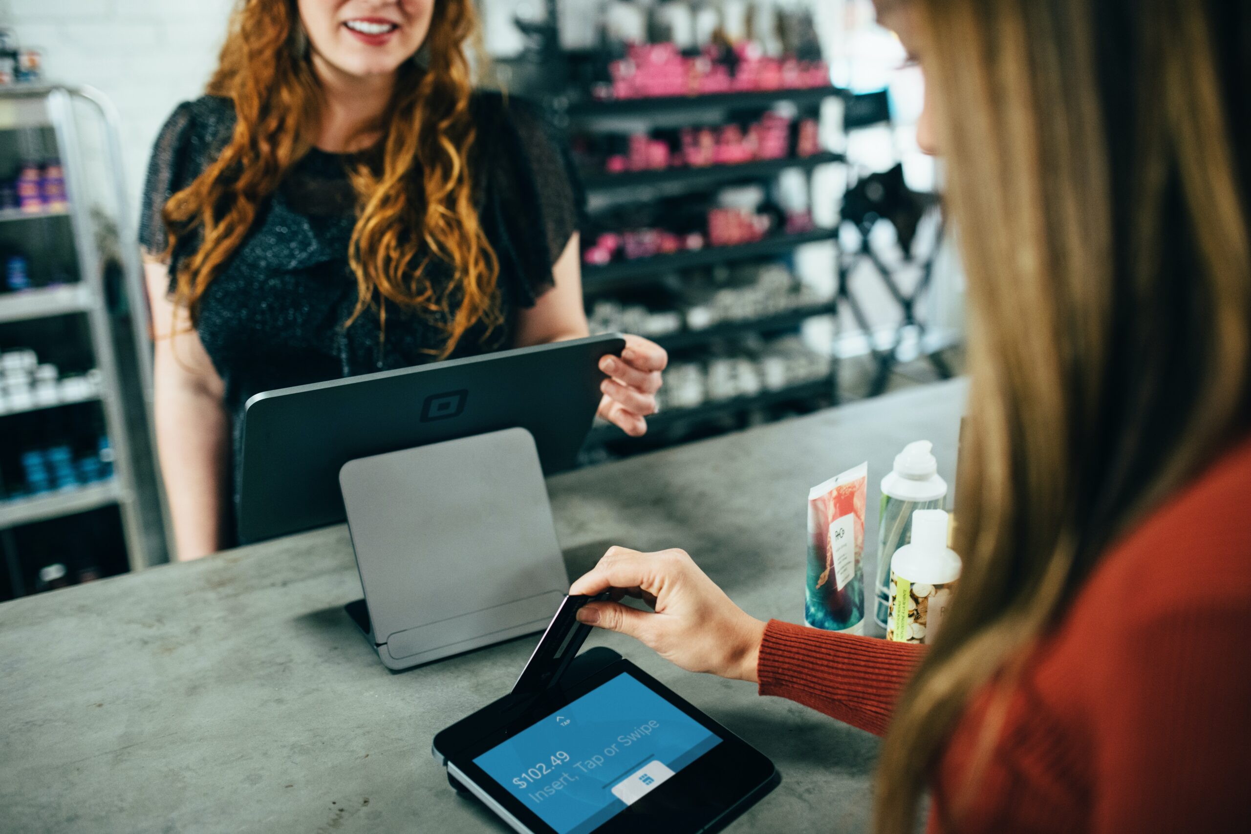 A woman inserts her credit card into a card reader. On the counter in front of her are several items she is purchasing, and the cashier watches from behind the counter with a smile.