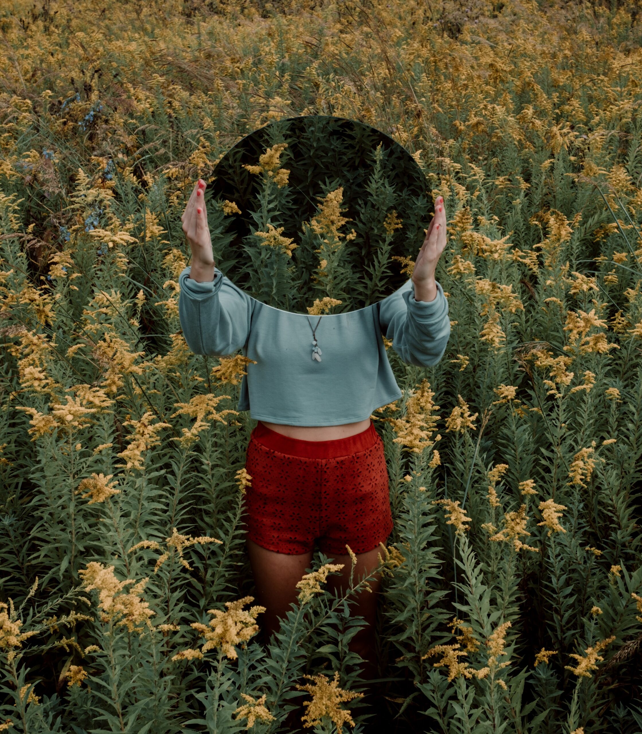 In a field of flowers, a woman stands holding a mirror over her face. It obscures her face, instead reflecting the flowers.