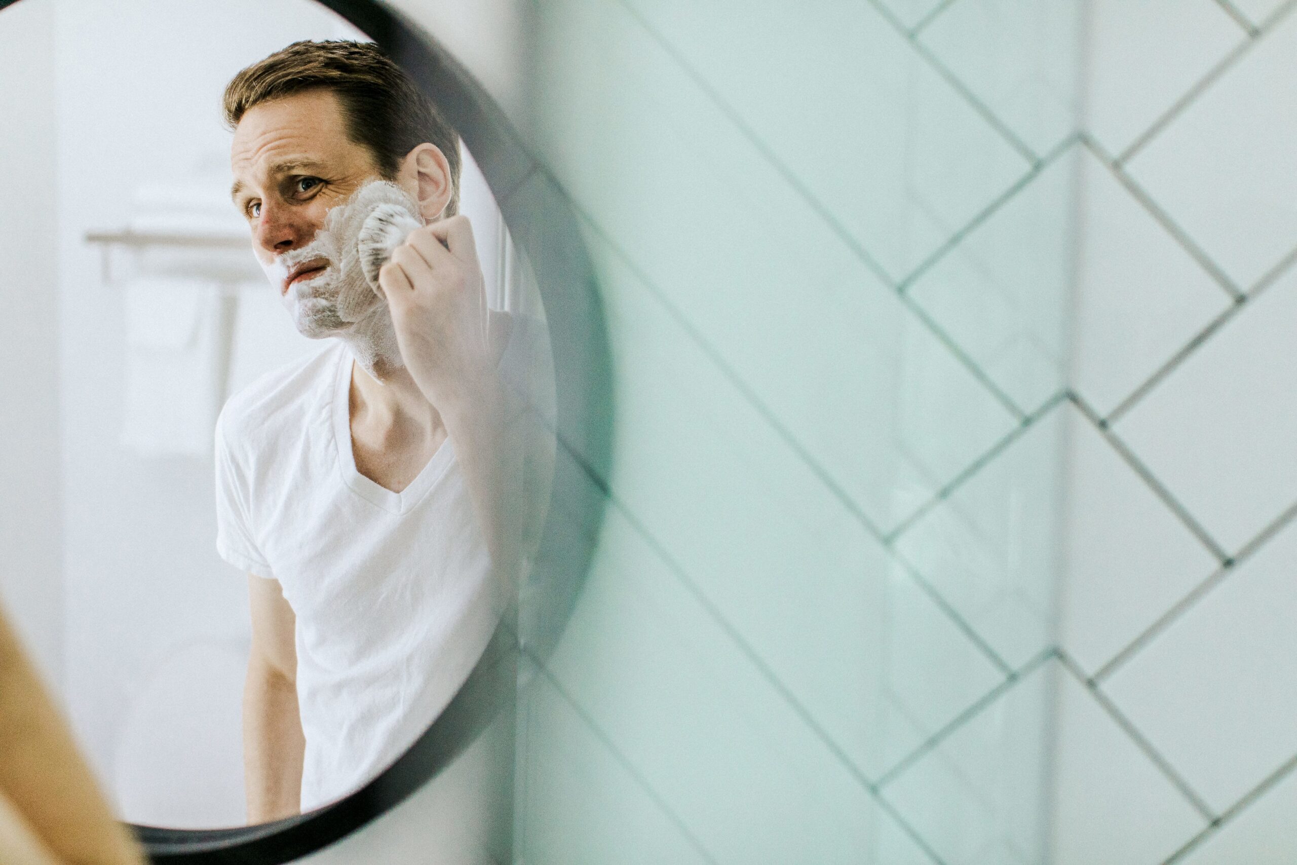 Man looking in mirror while rubbing shaving cream on his face.