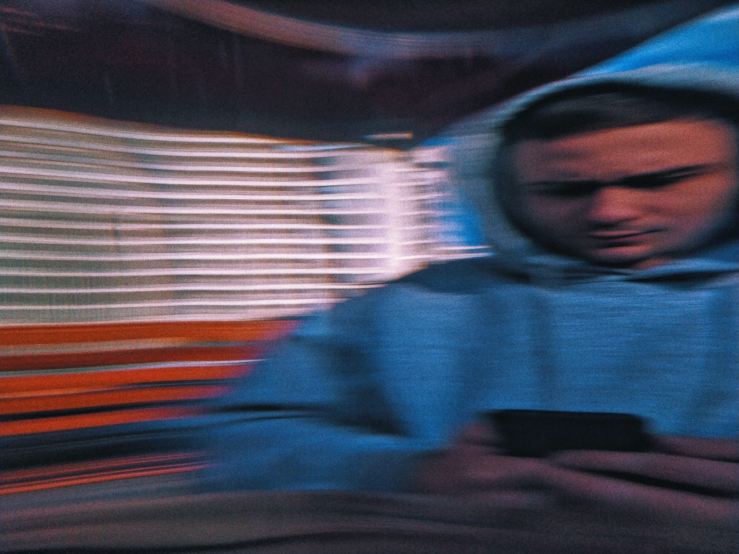 A motion blurred image of a man in a blue hoodie looking down at his phone in his hands. His brow is furrowed slightly.