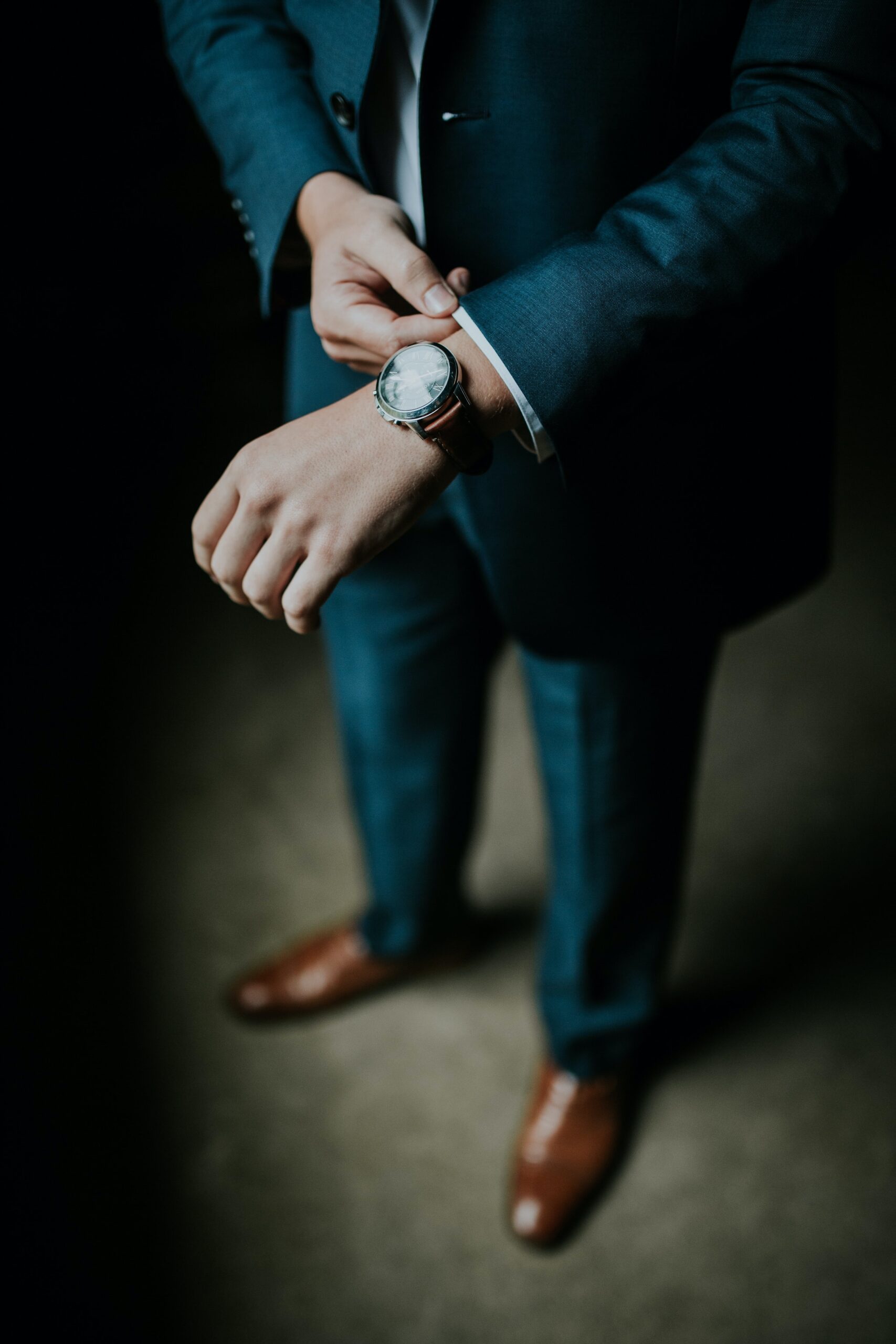 A view of a man in a blue suit from elbows down. He is wearing brown shoes and a watch, He is adjusting his left sleeve.