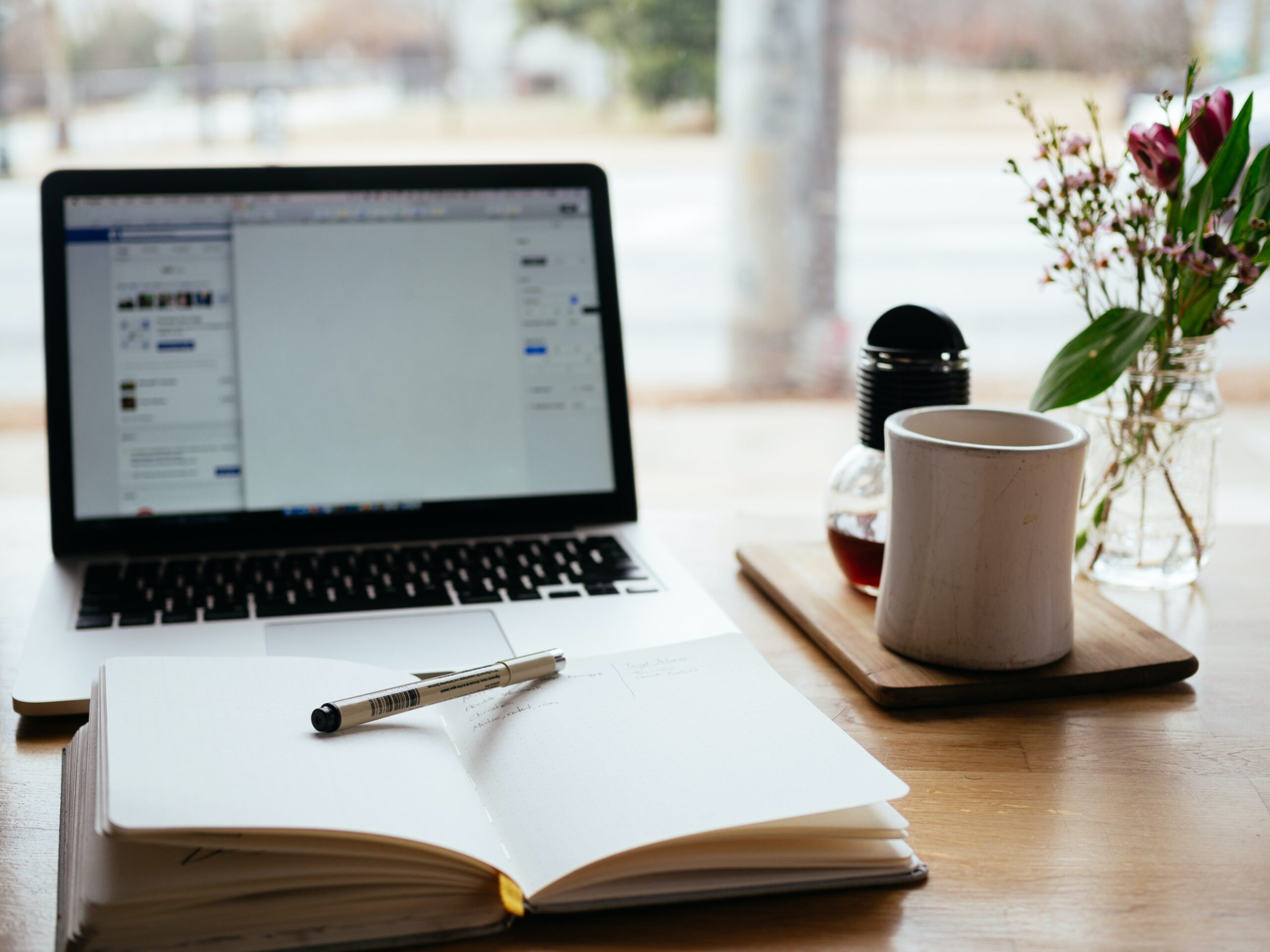 Laptop on desk with open notebook in front of it with a pen on top.