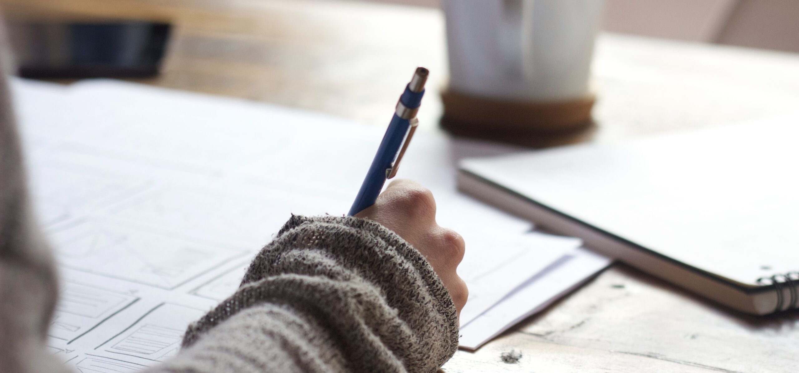 A Person writing on brown wooden table near white ceramic mug