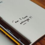 An notebook open to a page that is completely blank, aside from the words "Am I good enough?" written in black marker towards the bottom of the page. There is a pen and pencil resting in the spine of the notebook.