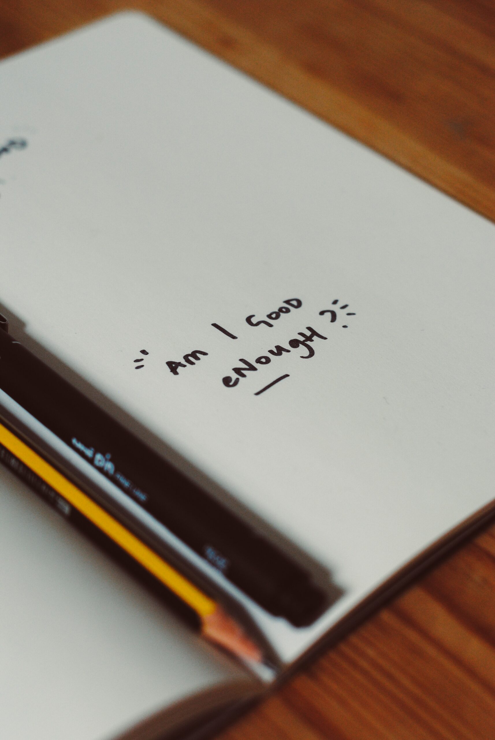 An notebook open to a page that is completely blank, aside from the words "Am I good enough?" written in black marker towards the bottom of the page. There is a pen and pencil resting in the spine of the notebook.