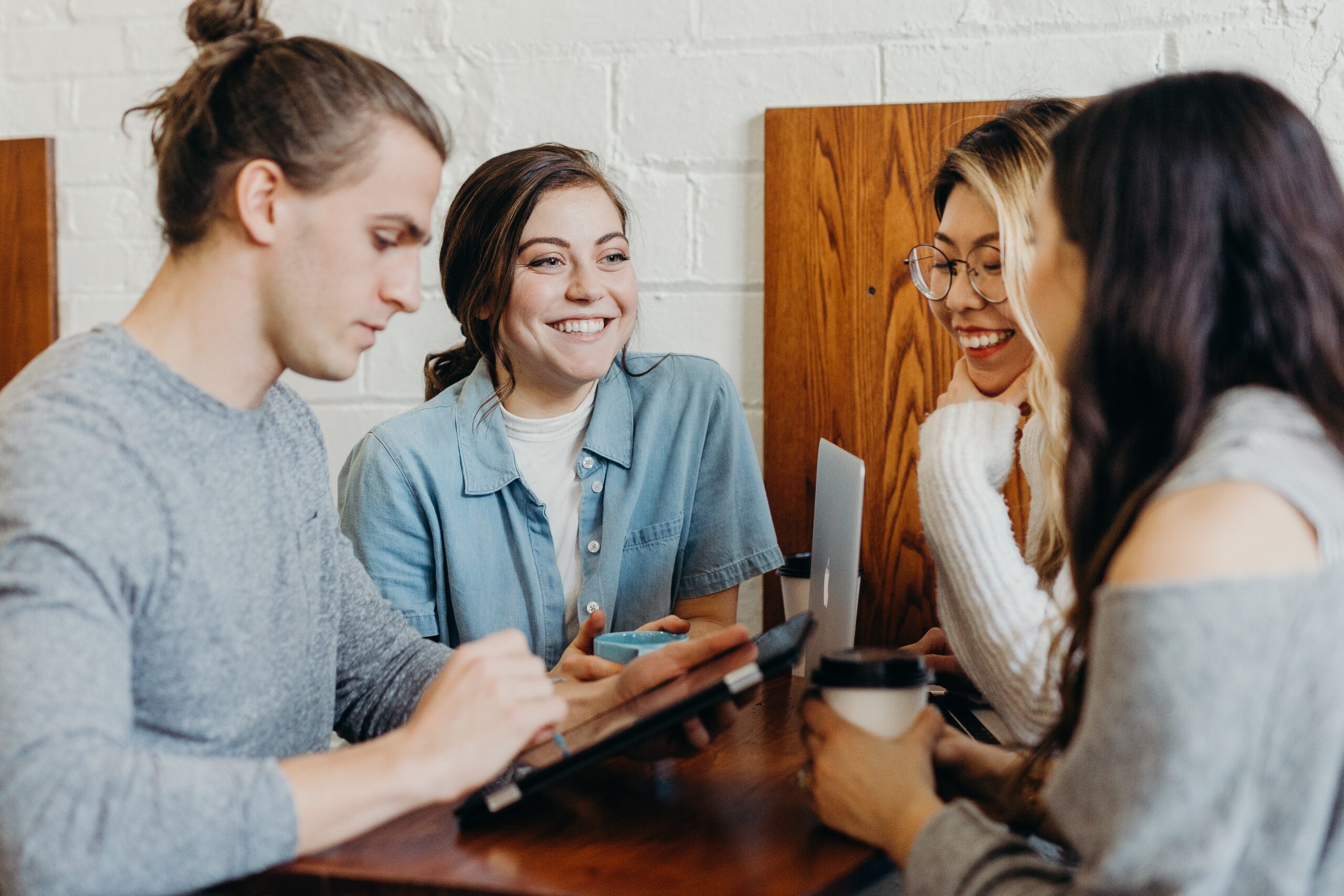 people sitting at a table, discussing things while having coffee. One of them has a computer, another one a tablet, and the other two are talking to the group