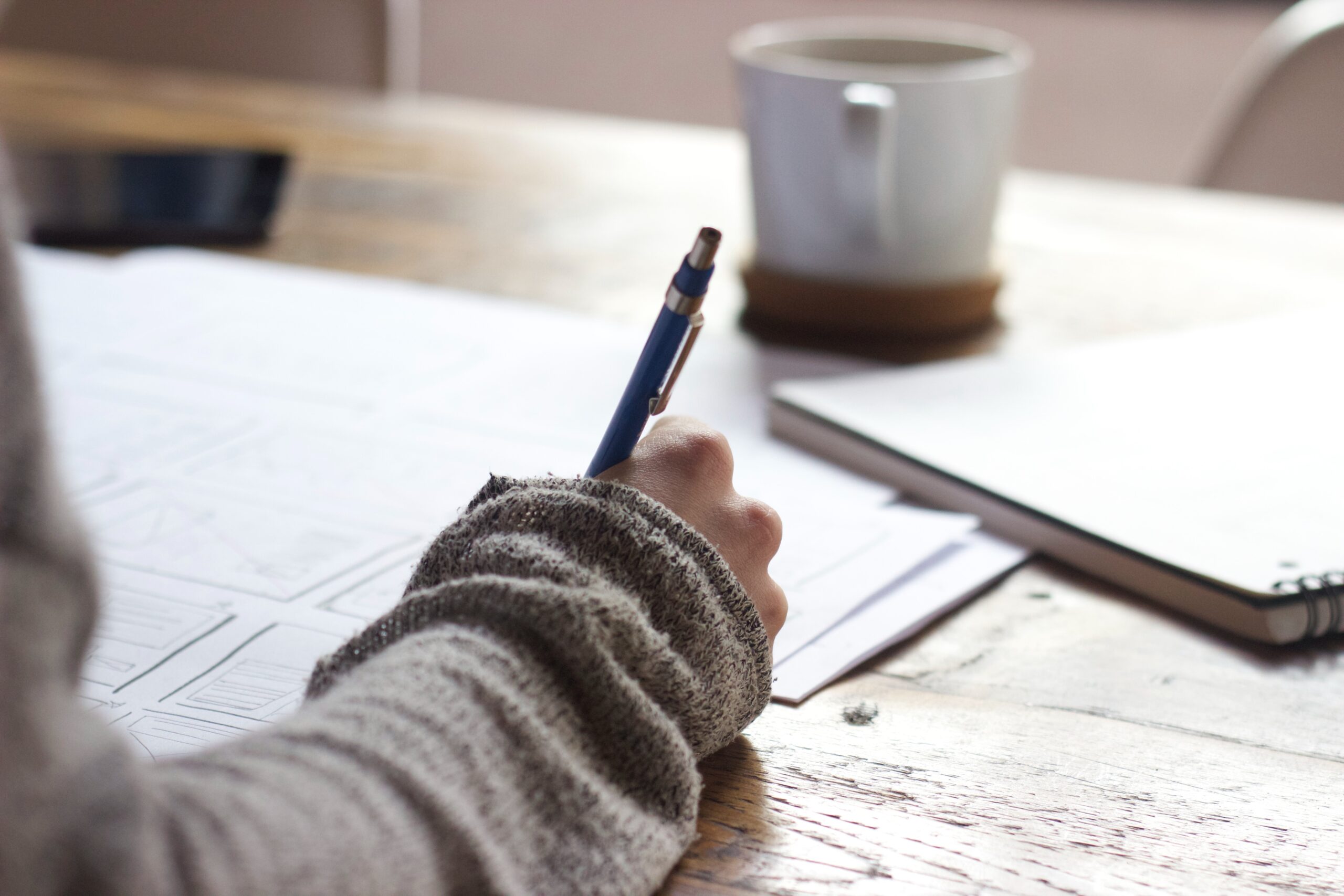 A person is writing on a piece of paper. They are surrounded by many different papers and they have a cup of coffee on the desk.