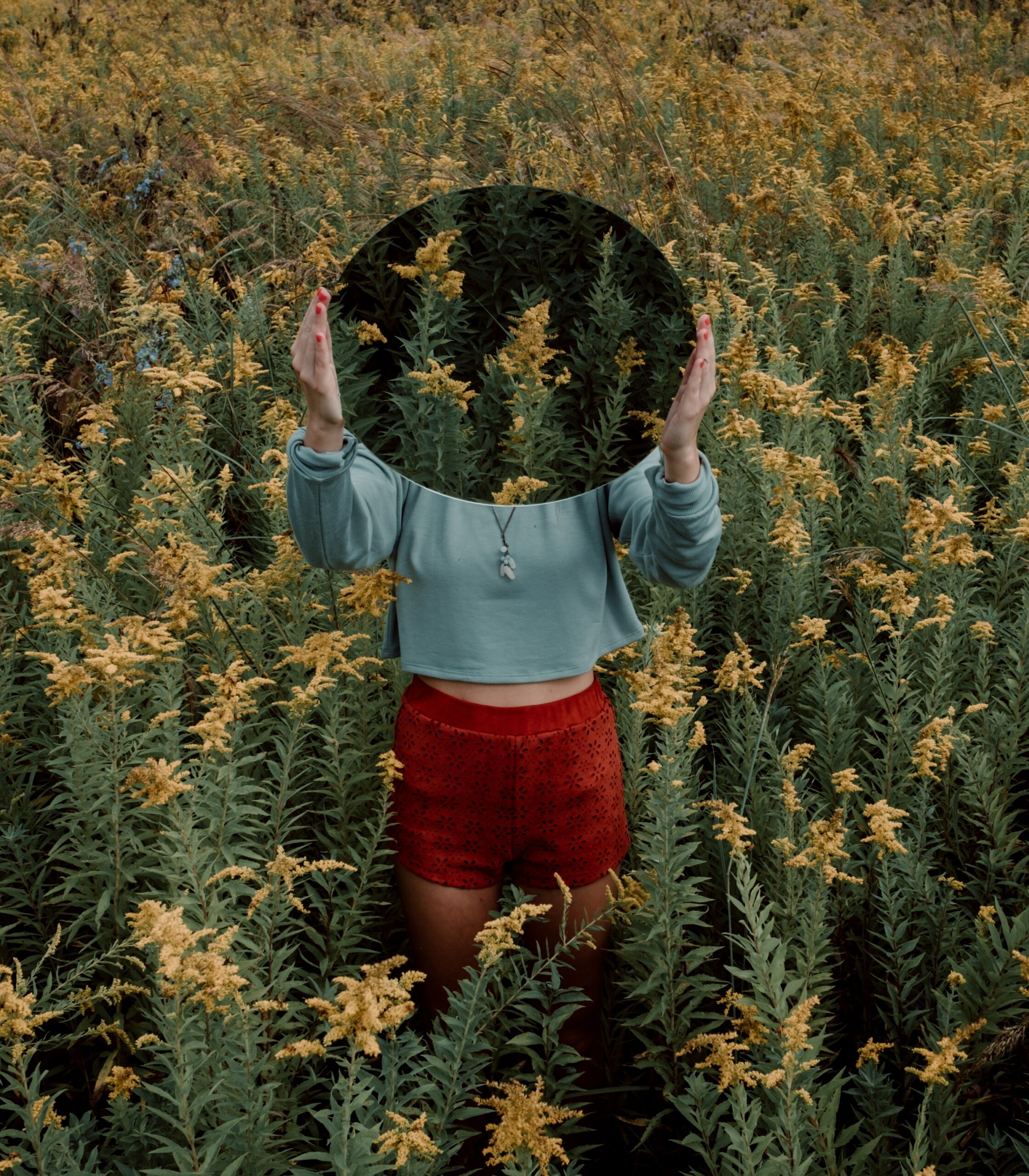 image of woman standing in a field of flowers covering her face with a mirror