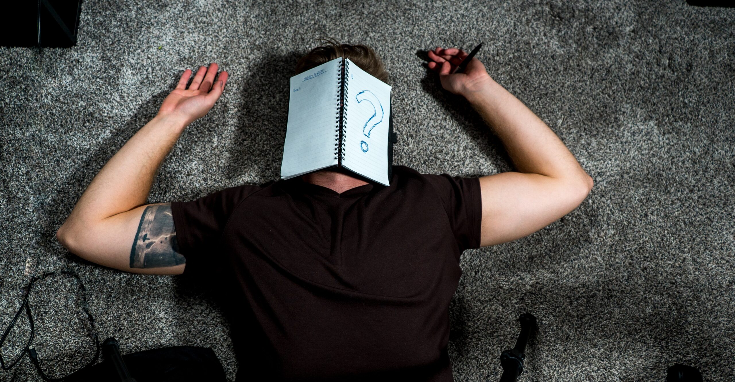 An individual with a black tee on the ground with a notepad covering his face. On the blank notepad, he has a question mark on it.