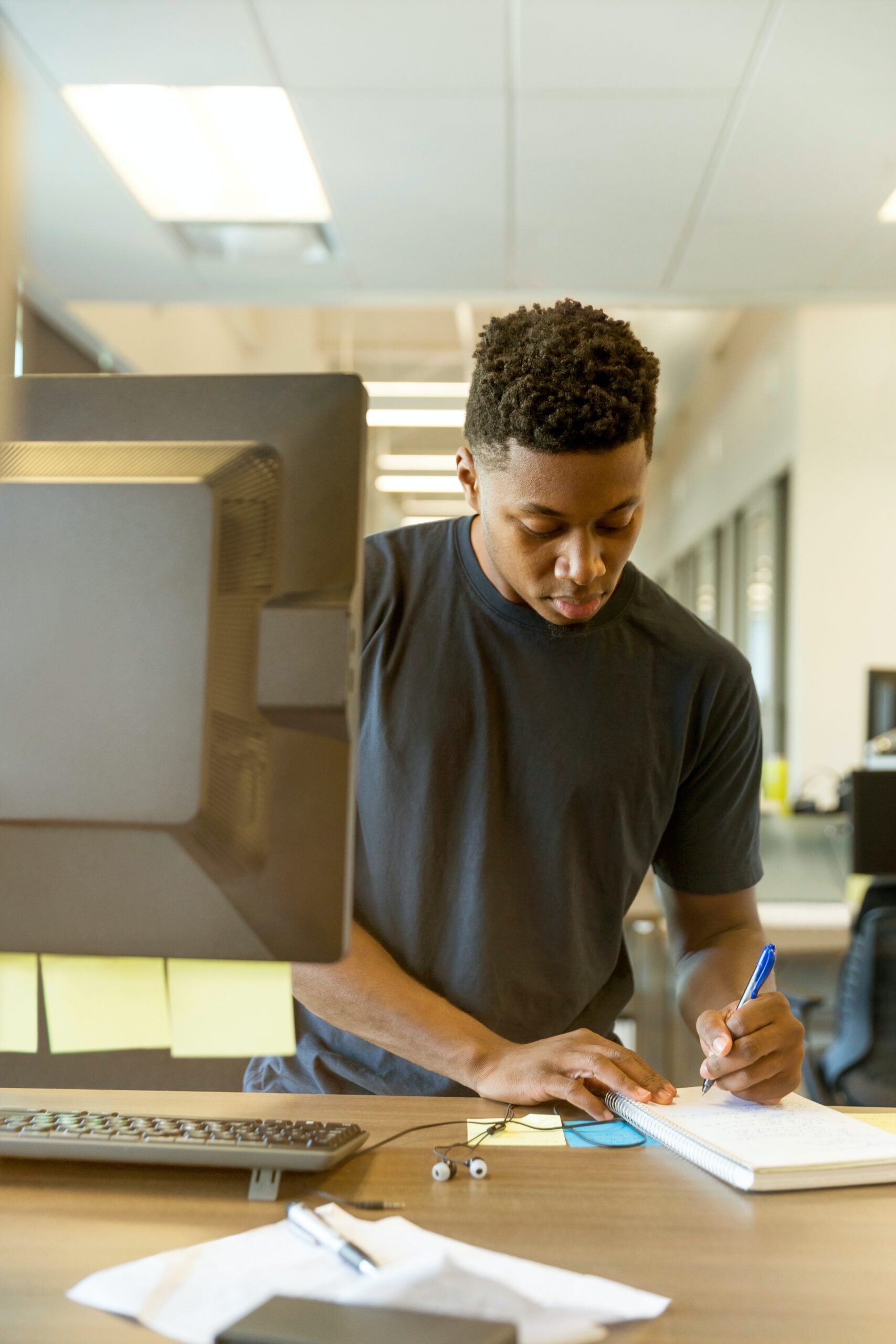 A man is standing in front of a computer and he's writing on a pad of paper. He has post-it notes around him and other pieces of paper.