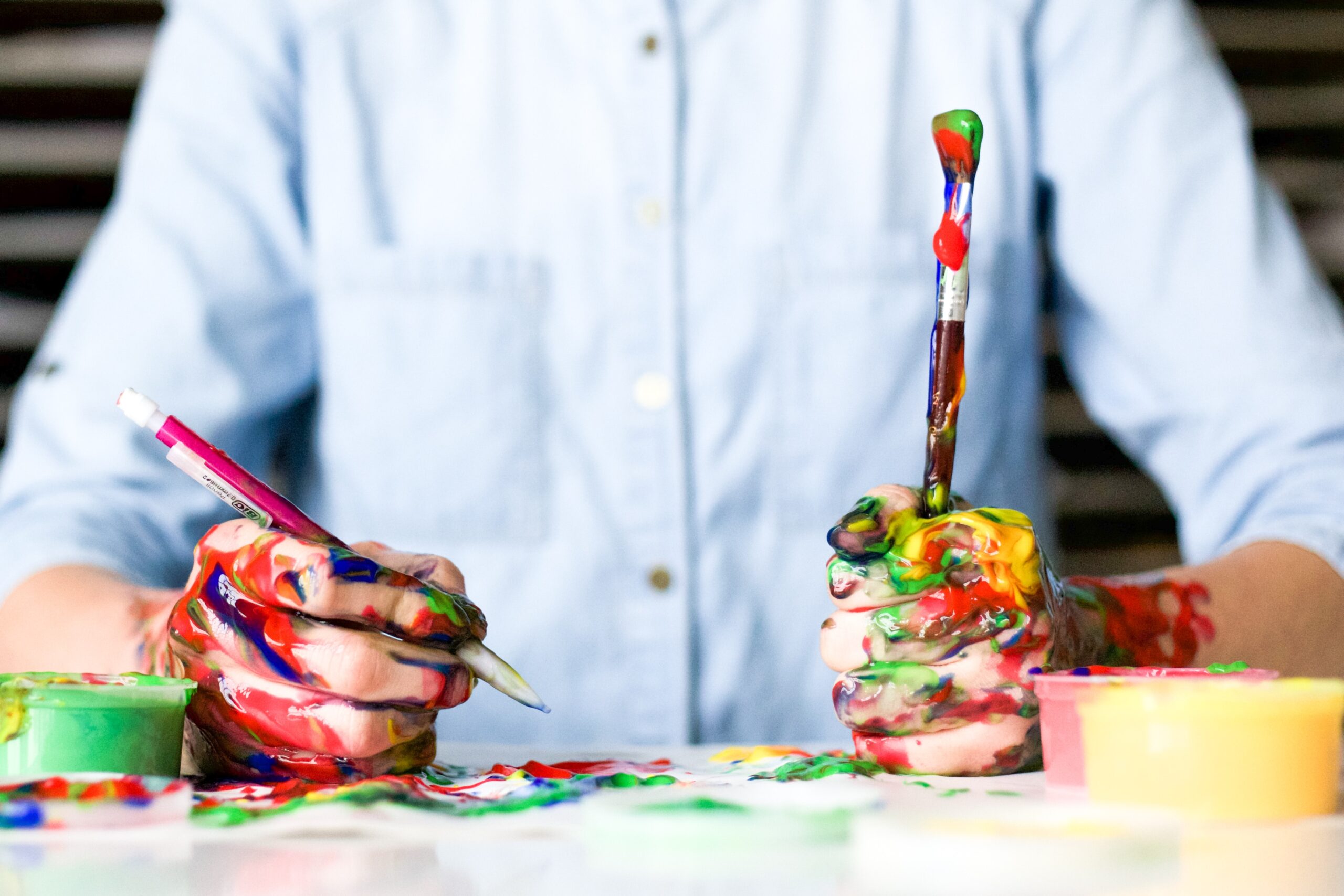 person's hands covered in paint while holding a paint brush in one hand and a retractable pencil in the other