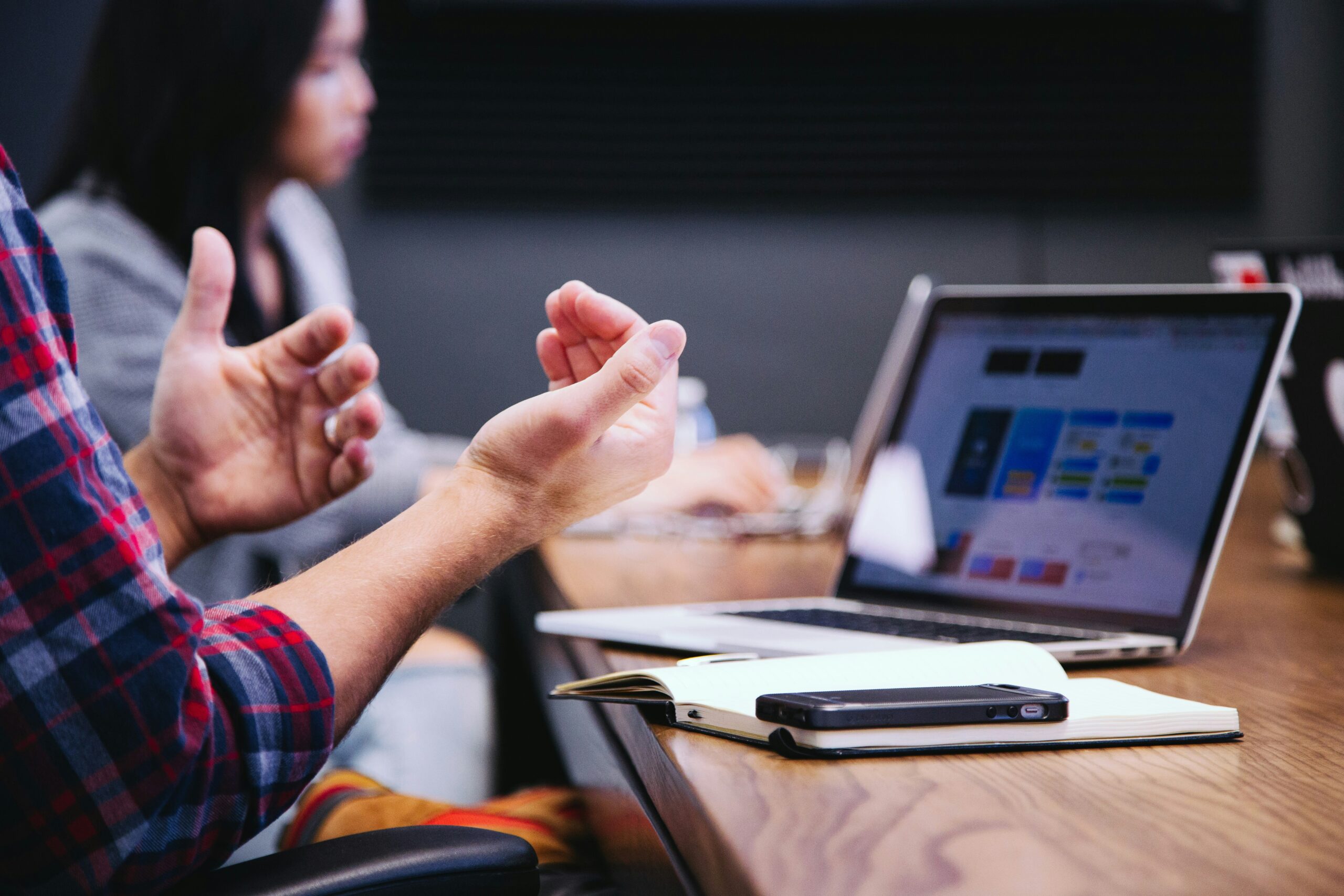 A person has their hands in front of them, using them to communicate with another person. They also have their laptop open and a notebook with a pen on it.