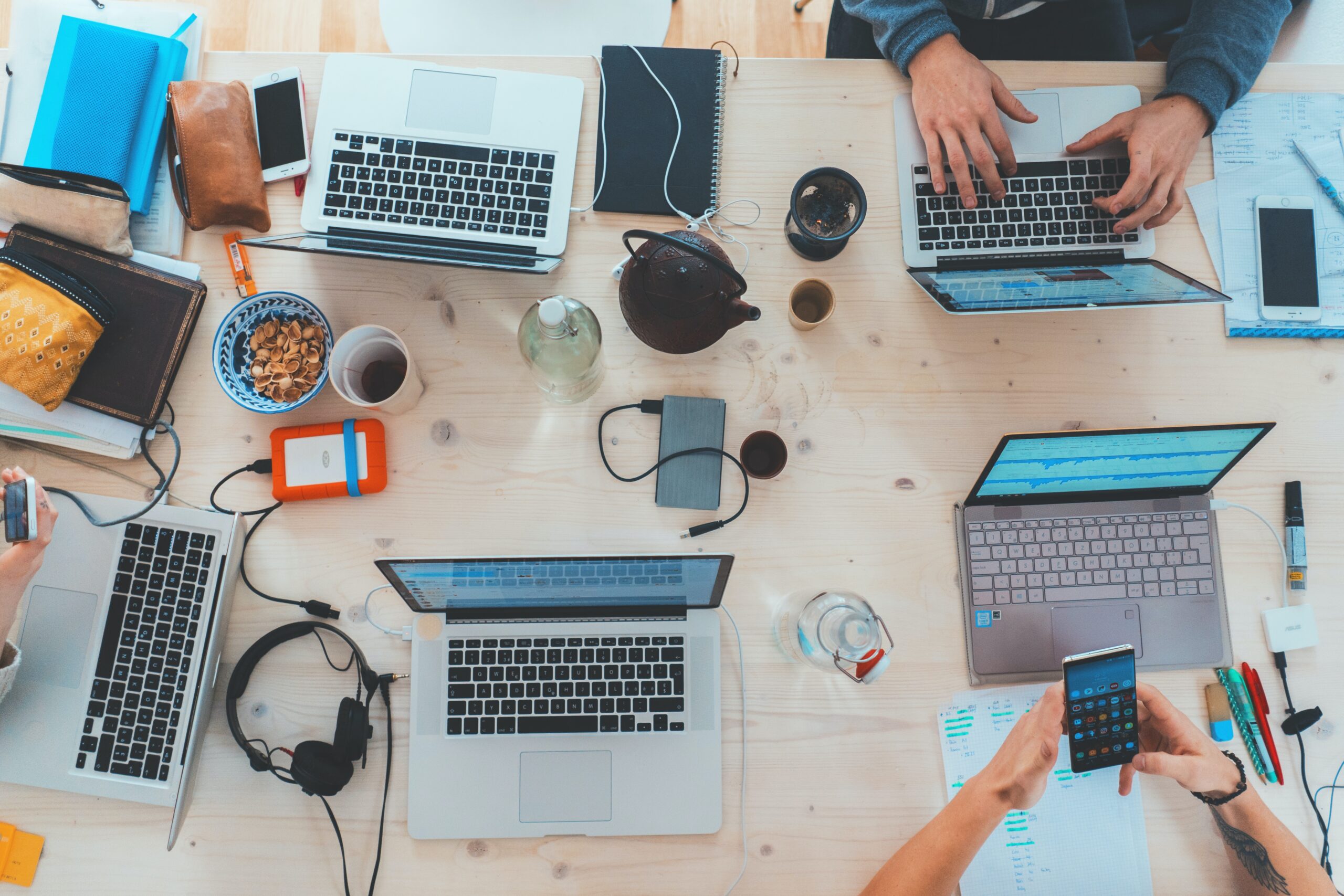 A tabletop filled with open laptops, headsets, and various coffee mugs. It represents a writer's downfall : technology.