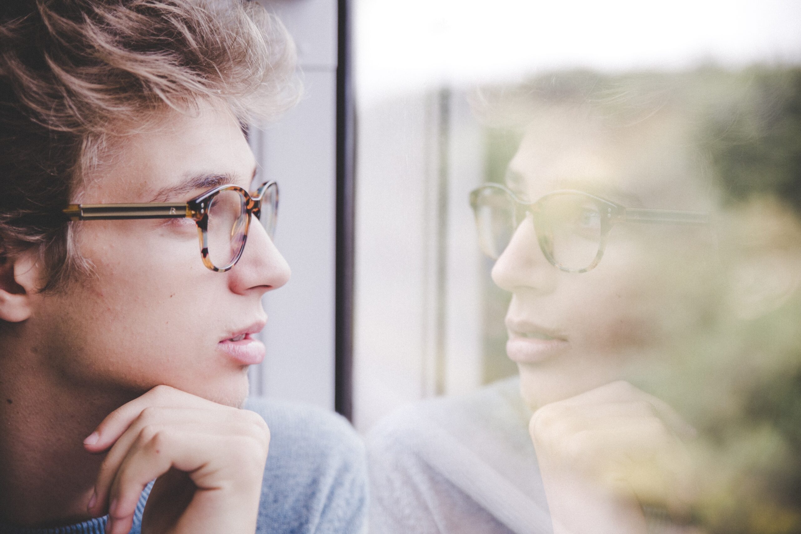 A person with glasses is looking at their reflection in the window.