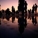 It is an image of a large group of people (silhouetted) who are cheering and enjoying their time together while the sun sets, and they can see the reflections of themselves through the water that reaches to their ankles.