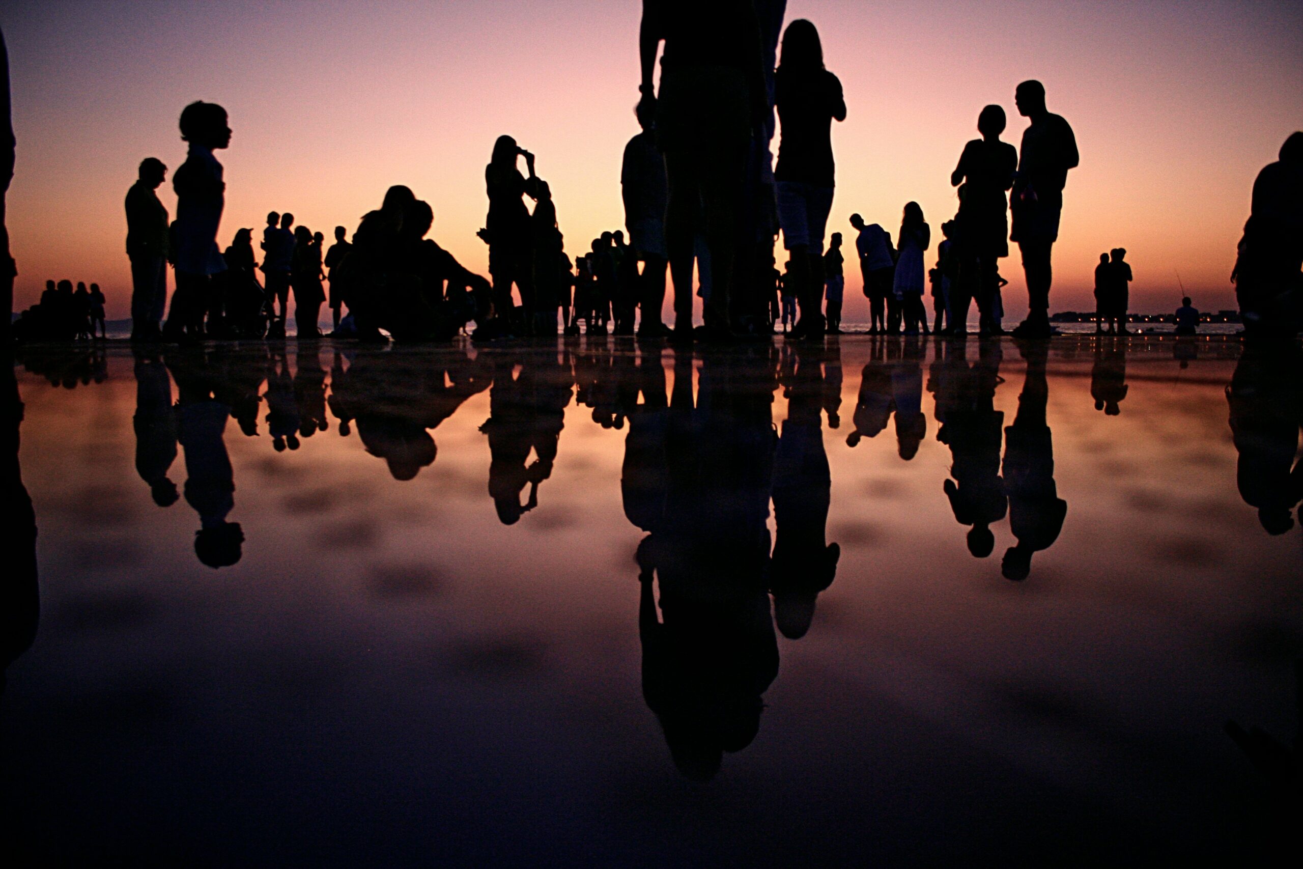 It is an image of a large group of people (silhouetted) who are cheering and enjoying their time together while the sun sets, and they can see the reflections of themselves through the water that reaches to their ankles.