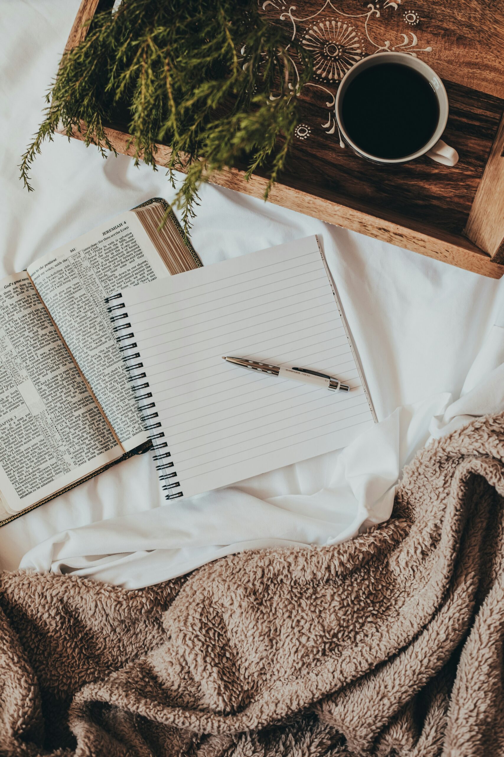 A tray with a cup of tea sitting on a bed. While a notebook with a pen sitting on top is laying on top of a book next to it.