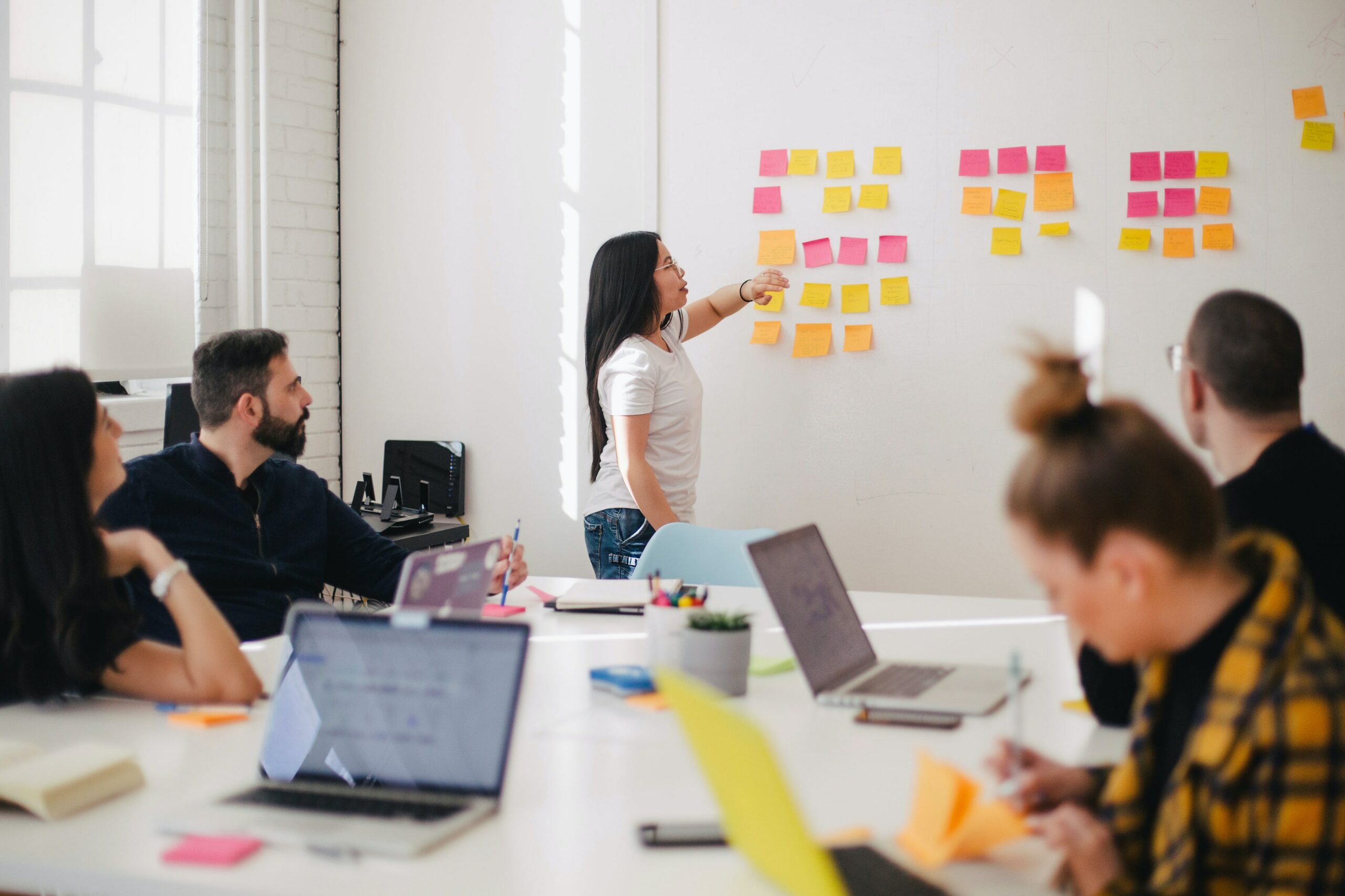 Everyone from the table is paying attention to a woman pointing at sticky notes who seems to have authority over the group