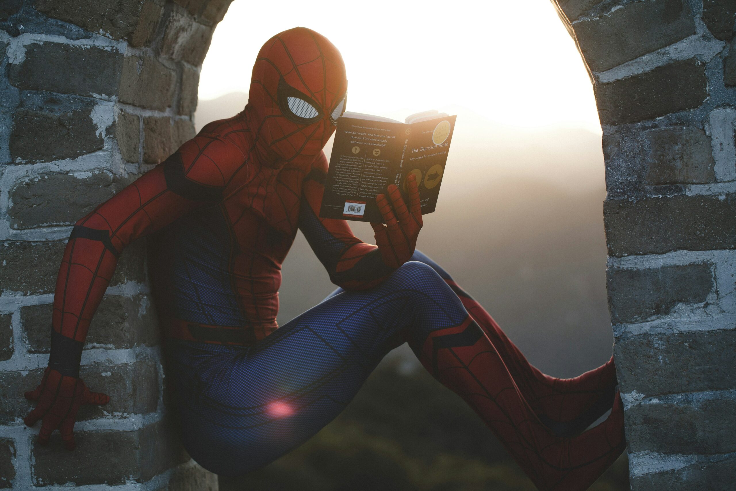 Spider-Man reading a book as he's sticking to a brick wall while the sun is bright.