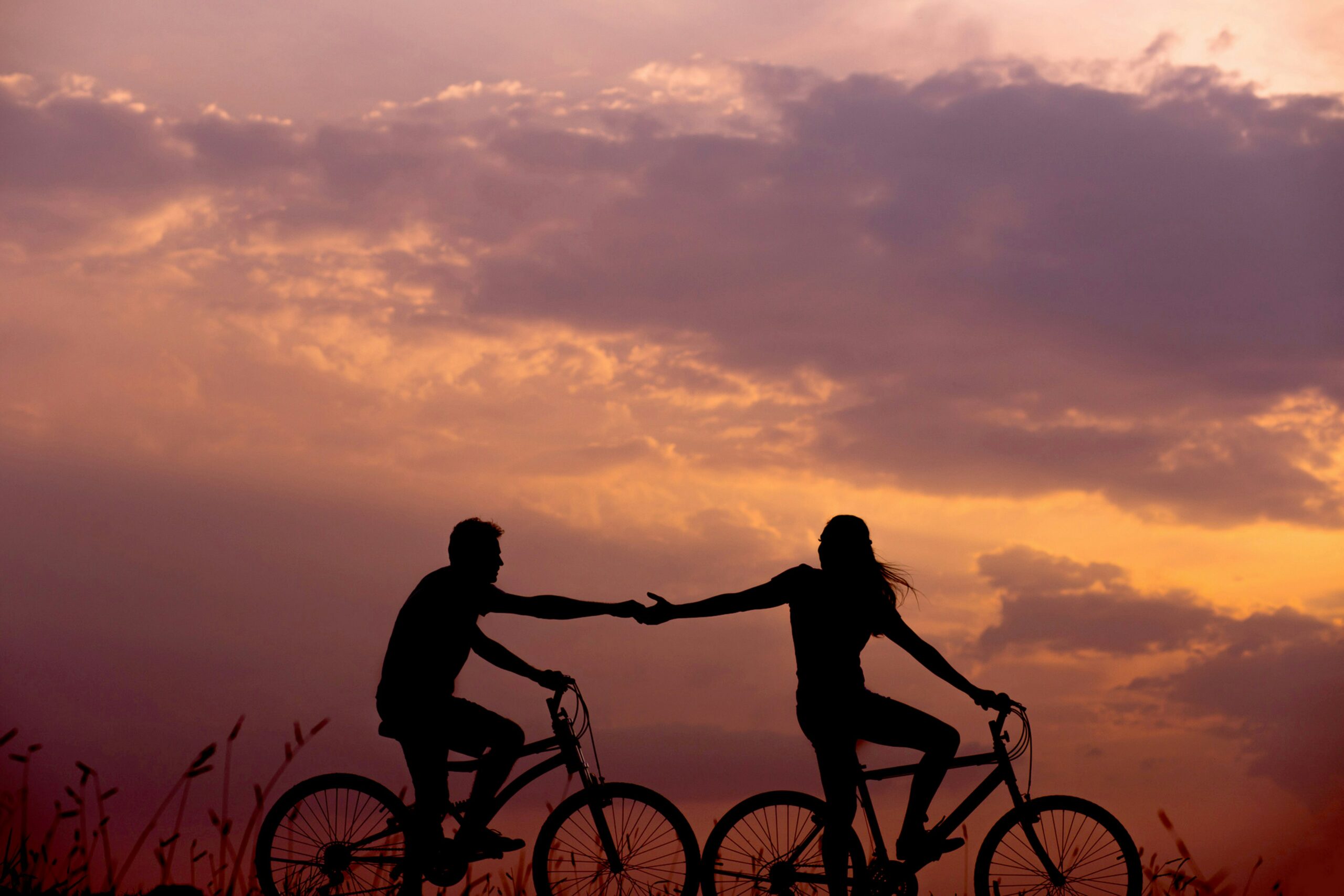 The silhouette of a man and woman holding hands while riding their bikes during a sunset.