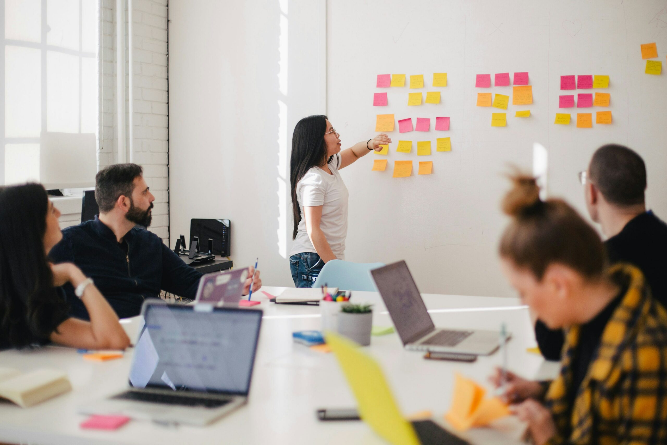 A group of men and women sitting at a table with their laptops out watching a woman explain something while she is pointing at her post it notes that are on the wall.