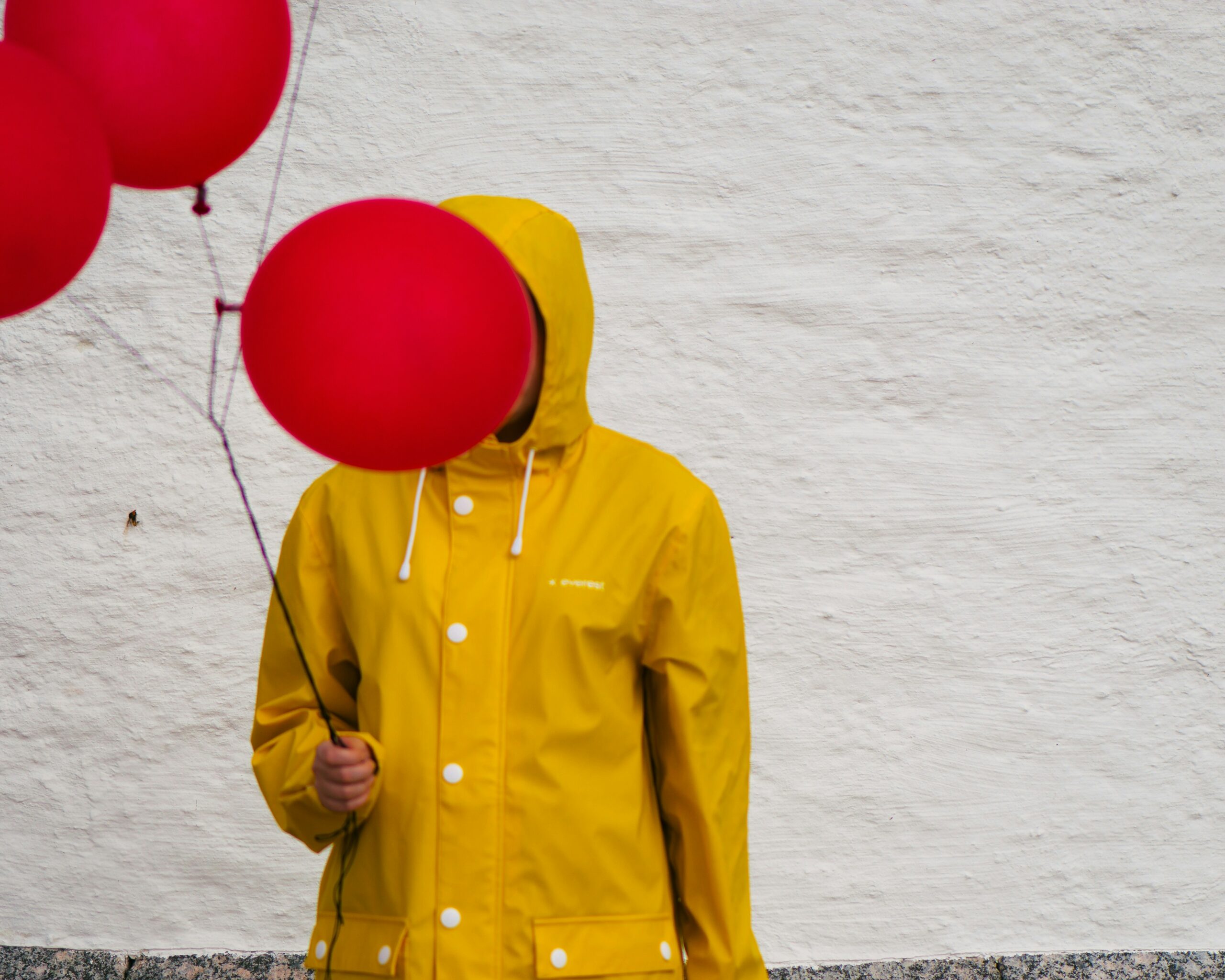 A person in a yellow raincoat holding a red balloon over their face.