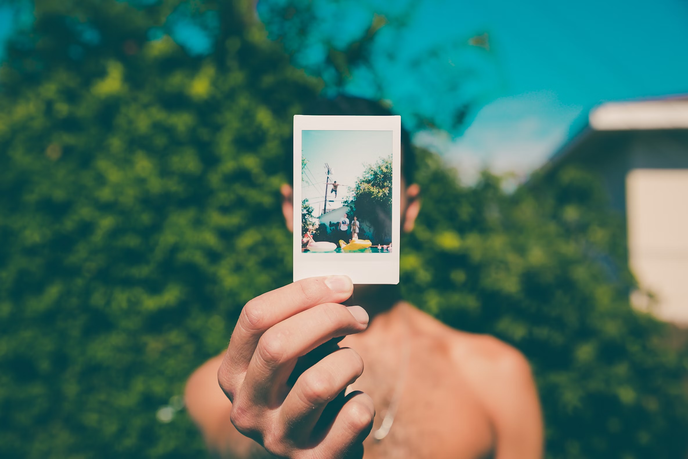 A topless man is holding a polaroid in the way of his face, close to the camera. Only the photo and his hand is in focus; the rest of his body and the thick bushes in the background are blurred.