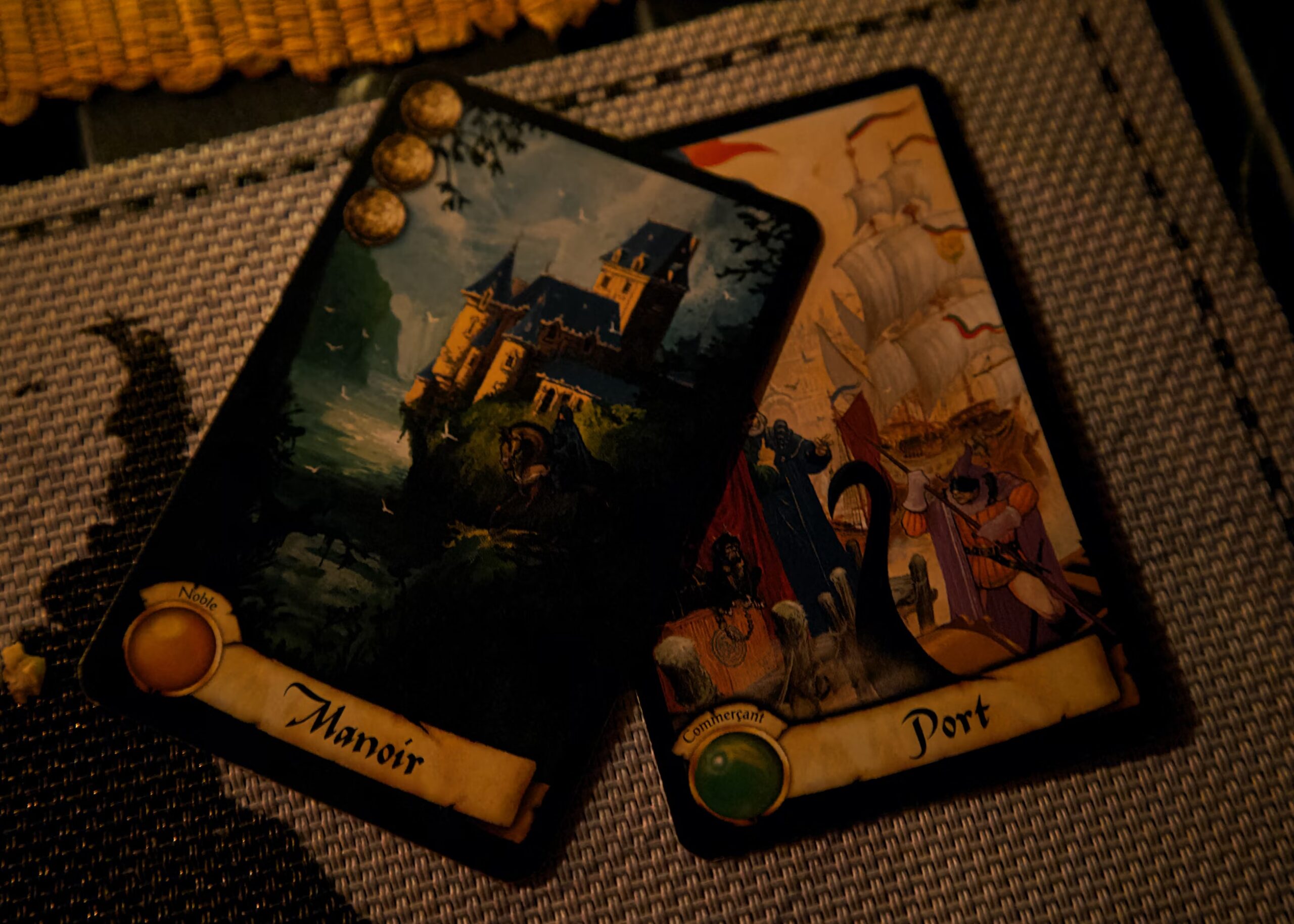 Up close, two cards lay on a mat in an orange-y, warm, and library-like room. One card reads “manoir” with the drawing of a castle atop a cliff, and the other one reads “port,” its drawing illustrating a bustling port during the medieval times.