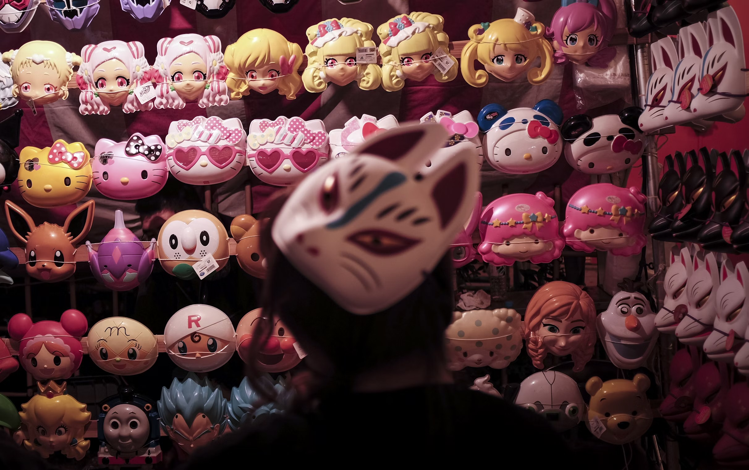 A person is surrounded by masks of characters from shows, such as Frozen, Pokemon, and Hello Kitty. The masks are hung up on the wall, and the room is very dimly lit, with a pink-ish haze to it.