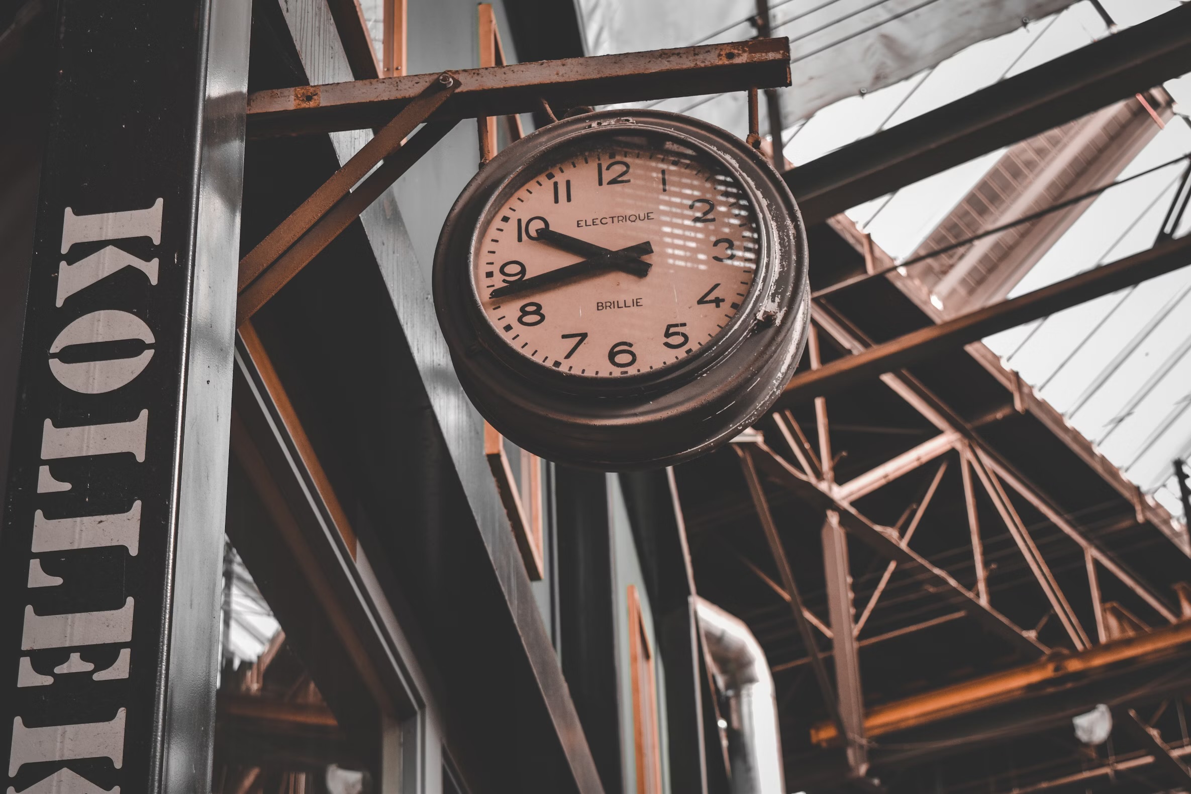 A clock hangs in what looks the outside of a cafe-like area. The vibe of the buildings is very rustic; the whole place has an orange and brown hue to it.