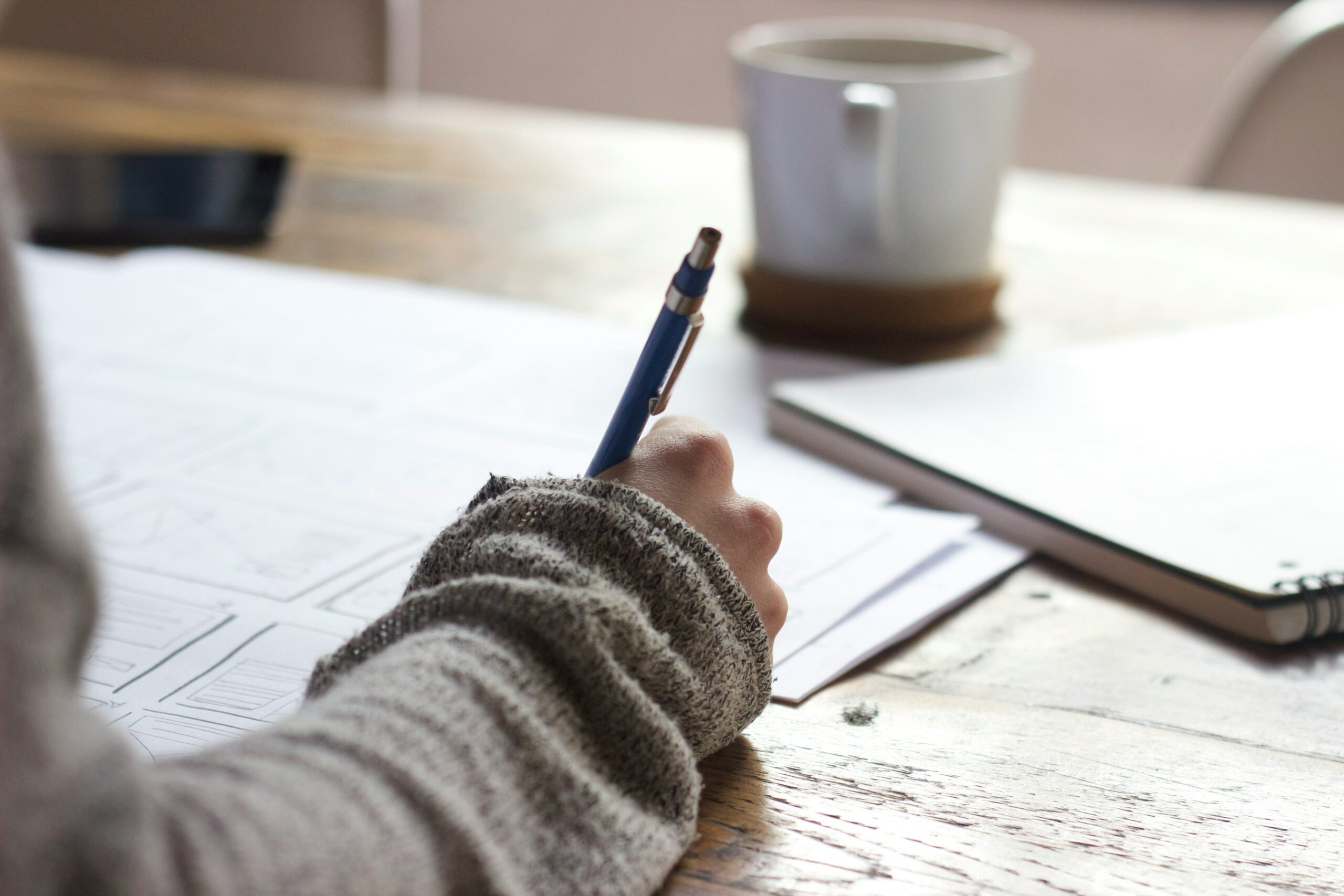 Picture of a sleeved arm and hand writing on paper, scattered on a desk.