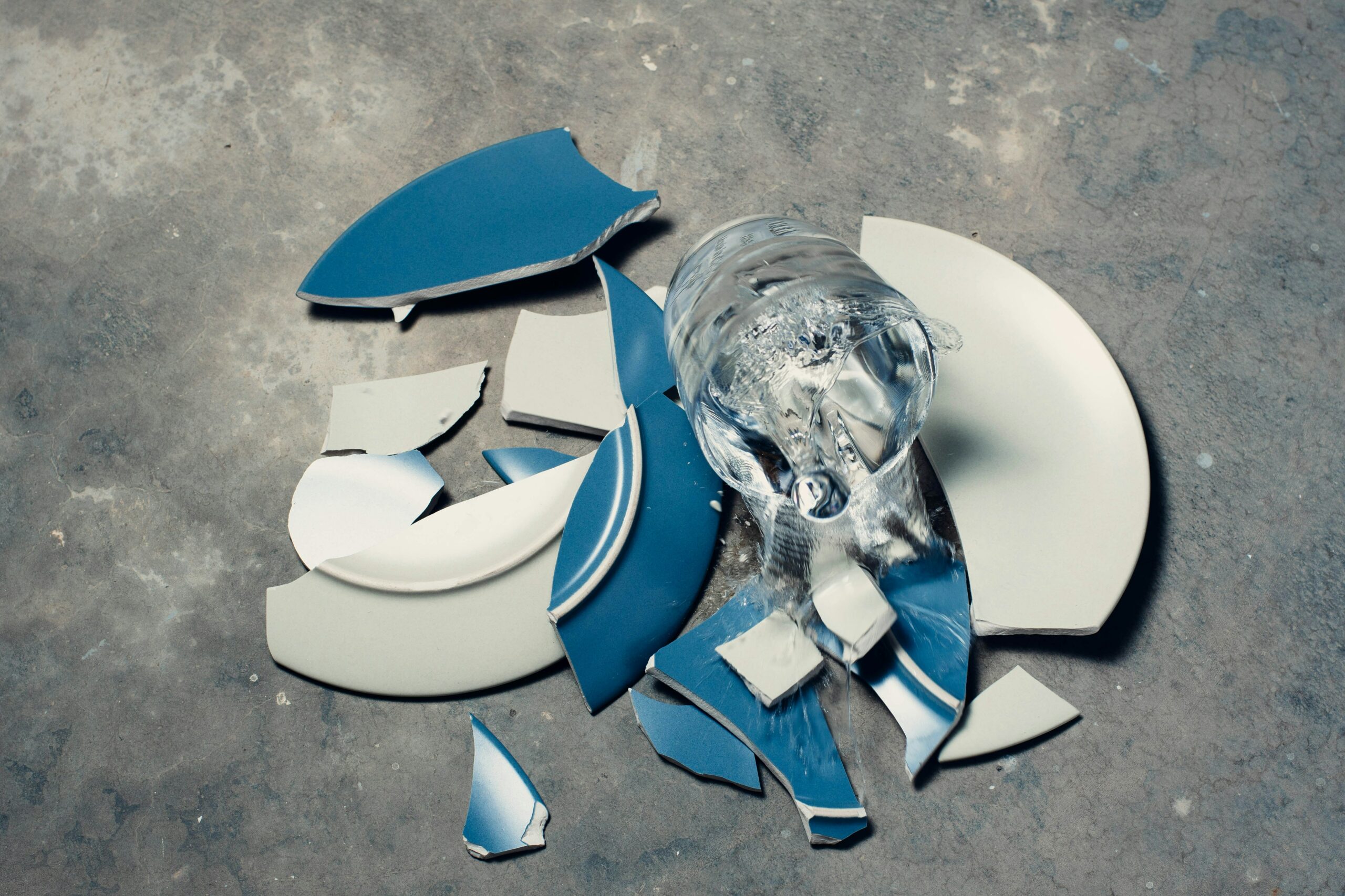 The shards of a blue plate are scattered atop a gray, marble counter. A glass full of water is mid-fall above the broken plate.