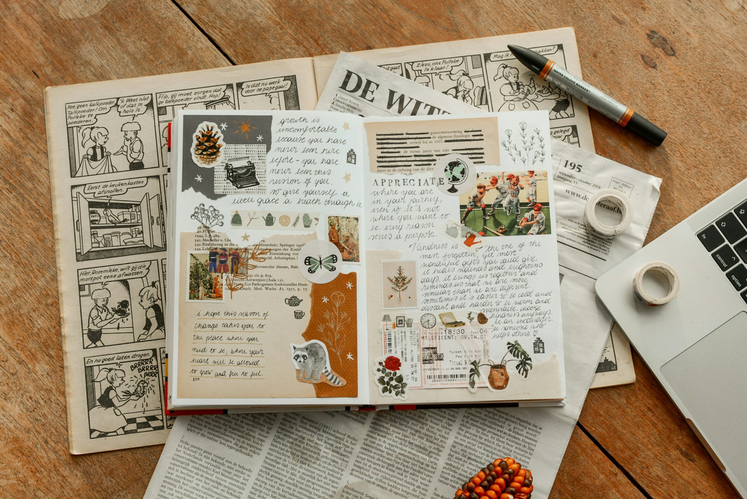 A close up of an open journal atop a comic and magazine. There is a pen and tape scattered around the sides of the photo. Inside the journal, there are a bunch of stickers, photos, and different kinds of paper decorating it.